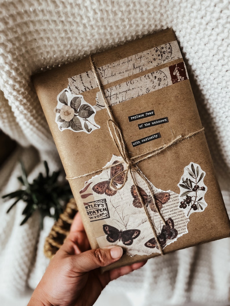May include: A brown paper package wrapped with twine and decorated with vintage stamps, paper scraps, and floral stickers. The package has a handwritten message that reads "replace fear of the unknown with curiosity".