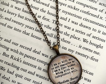 May include: A bronze chain necklace with a round glass pendant featuring a quote from Charlotte Bronte's Jane Eyre: "I am no bird, and no net ensnares me. I am a free human being with an independent will." The pendant is on a book with the text "...it owns me."