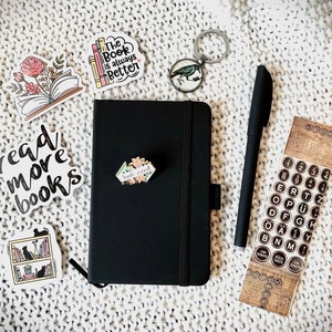 May include: A black notebook with a book-themed sticker on the front. The notebook is surrounded by other stickers, including one that says "read more books" and another that says "The Book is always Better". There is also a pen, a key chain with a bird, and a sheet of typewriter key stickers.