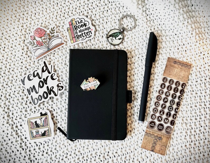 May include: A black notebook with a book-themed sticker on the front. The notebook is surrounded by other stickers, including one that says "read more books" and another that says "The Book is always Better". There is also a pen, a key chain with a bird, and a sheet of typewriter key stickers.