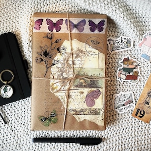 May include: A brown paper-wrapped journal with decorative butterfly and floral stickers, a black pen, and several other stickers with book-themed designs. The stickers include "Book Hoarder", "Easily Distracted by Books", and a stack of books.