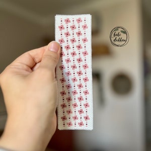 May include: A pink and white cross-stitch bookmark with a geometric pattern. The bookmark is handmade and features a simple design.