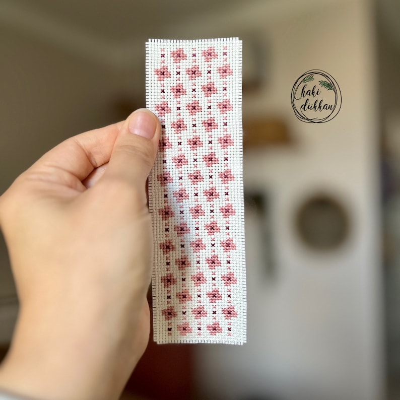May include: A pink and white cross-stitch bookmark with a geometric pattern. The bookmark is handmade and features a simple design.