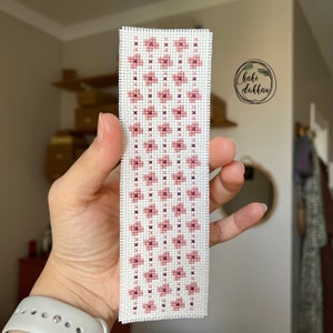 May include: A white bookmark with a pink and burgundy floral pattern. The bookmark is hand-stitched and has a white border. The text "haki d&uuml;kkan" is visible in the background.