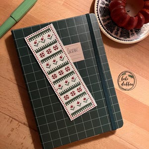 Pode incluir: Um caderno verde com um padrão de grade e um marcador de página branco com um design floral vermelho e verde. O marcador de página está etiquetado como "TURKAN".