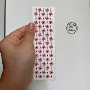 May include: A white bookmark with a pink floral pattern. The bookmark is made with cross-stitch embroidery.