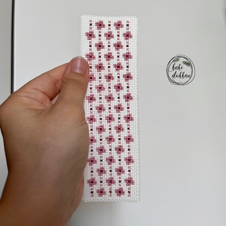 May include: A white bookmark with a pink floral pattern. The bookmark is made with cross-stitch embroidery.