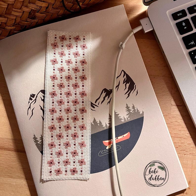 May include: A white bookmark with a pink and white cross-stitch pattern. The bookmark is on a notebook with a mountain scene and the text "Chaki Dukkan" in a circle.