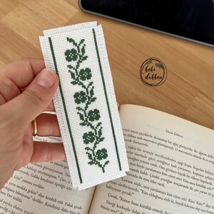 Puede incluir: Un marcapáginas blanco con un diseño floral bordado en verde. El marcapáginas está hecho a mano y presenta un diseño simple y elegante.