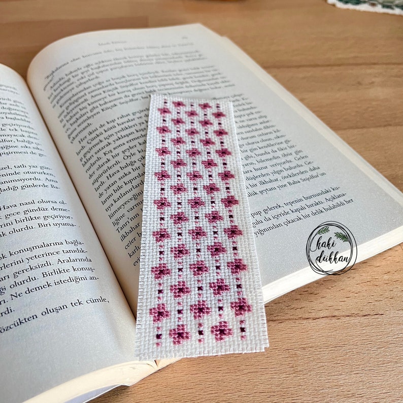 May include: A pink and white bookmark with a floral pattern. The bookmark is made of fabric and has a white background with pink flowers.