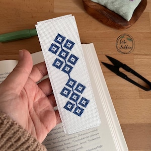 Pode incluir: Um marcador de página branco com um padrão geométrico azul e branco. O marcador de página é feito à mão e apresenta um design simples.