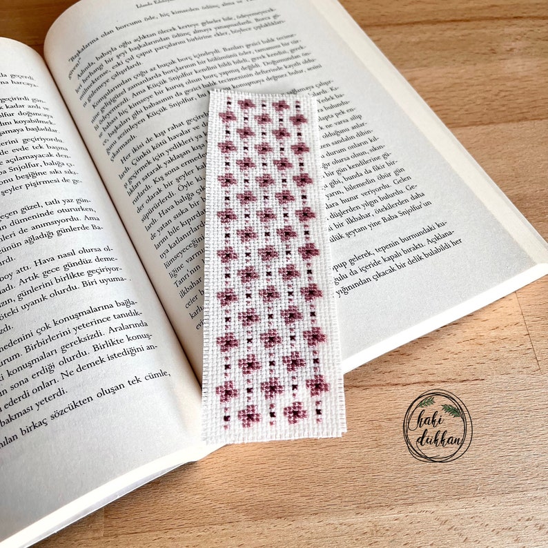 May include: A close-up of a pink and white embroidered bookmark with a floral pattern. The bookmark is lying on top of an open book.