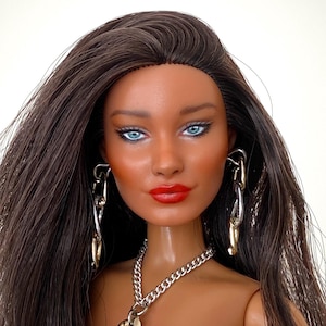 May include: A doll with long dark brown hair, bright blue eyes, and vibrant red lipstick. The doll wears silver and gold-toned chain link earrings and a matching chain link necklace with a large circular pendant against a black top.