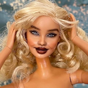May include: A doll with long, wavy blonde hair, blue eyes, and dark brown lipstick. The doll has a light skin tone and is wearing a bright pink textured garment. The facial features include defined eyebrows, dark eyeliner, and a subtle smile.