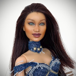May include: A doll with long dark brown hair, light skin, and green eyes. The doll wears a dark blue denim choker with three small gold-toned circular studs and a matching off-the-shoulder denim top featuring frayed edges and gold trim.