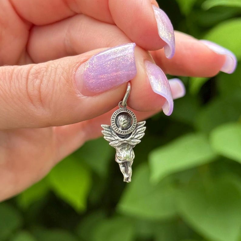 May include: A silver-toned angel charm with detailed wings and a halo. The charm is suspended from a small hoop, suitable for a necklace or bracelet. The angel figure is intricately designed, showcasing a classic, celestial theme.