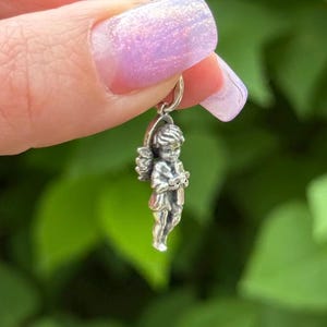 May include: A small, silver-colored angel pendant with detailed wings and clothing. The angel is holding a small object, possibly a book or scroll. The pendant is suspended from a silver loop, suitable for a necklace or charm bracelet.