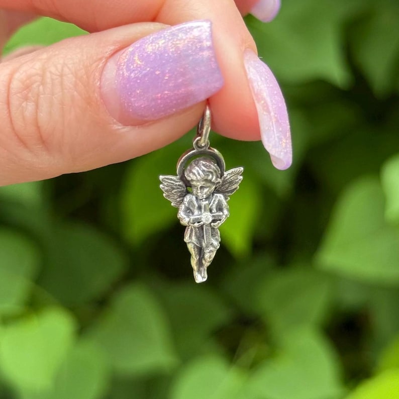 May include: A small, silver-colored angel pendant with detailed wings, halo, and clothing. The pendant is suspended from a small loop, suitable for a necklace or charm bracelet. The angel is holding a small object.
