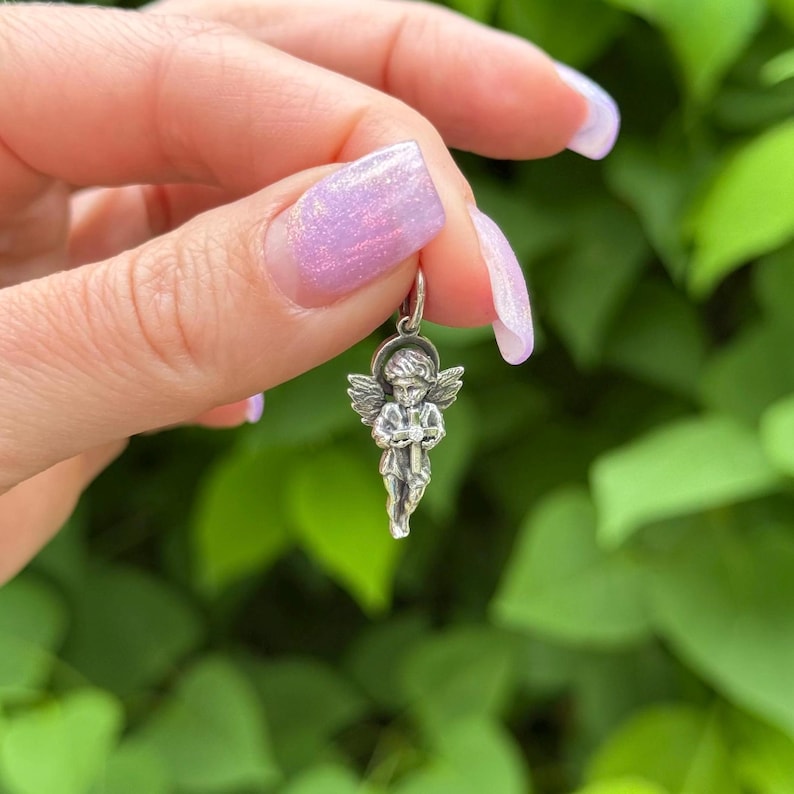 May include: A small, silver-colored angel charm with detailed wings and a halo. The charm is suspended from a small loop, suggesting it's designed for a necklace or bracelet. The angel is holding a small object, possibly a bow or a musical instrument.