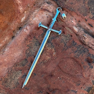 Peut inclure: Un pendentif en forme d'épée argentée avec un design détaillé. Le pendentif est posé sur une surface de roche rouge.