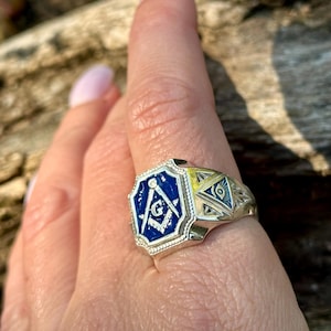 May include: A silver ring with a blue enamel Masonic symbol and a gold-toned all-seeing eye design. The ring is worn on a person's finger.