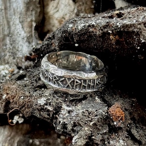 Può includere: Un anello d'argento con una superficie strutturata e simboli runici incisi. L'anello è posto su uno sfondo scuro e strutturato, evidenziando la sua lucentezza metallica e il suo design intricato. Il design dell'anello suggerisce un tema storico o fantasy.