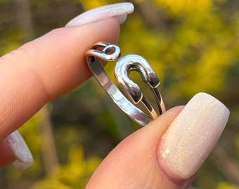 Pode incluir: Um anel de prata em forma de alfinete de segurança. O anel é usado em um dedo com esmalte de unha branco.