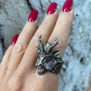 May include: A silver-toned ring featuring a detailed goat head design. The ring showcases the goat's horns, beard, and facial features. The ring is worn on a finger with red nail polish.