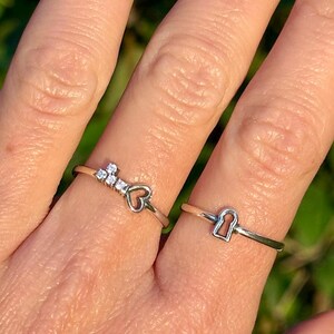 May include: Two silver rings are displayed. One ring features a key design with three small clear stones and a heart. The other ring has a keyhole design. The rings are on a finger with a blurred green background.