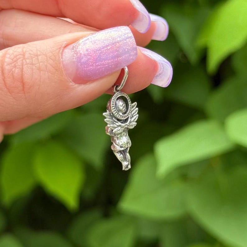 May include: A small, silver-colored cherub pendant with detailed wings and a halo. The cherub is holding a circular object. The pendant is suspended from a small loop, ready to be added to a necklace or bracelet. The background is blurred green foliage.