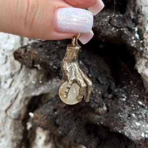 May include: A gold-colored pendant featuring a detailed hand holding a clear, round crystal ball. The hand is intricately sculpted, with visible fingers and a textured surface. The crystal ball appears to be translucent, reflecting light. The pendant is designed for jewelry.