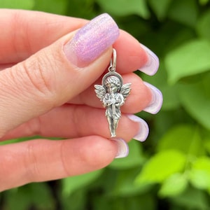 May include: A small, silver-colored angel pendant with detailed wings and a halo. The angel is holding a cross. The pendant is suspended from a small loop, ready to be added to a necklace or bracelet. The background is blurred green foliage.