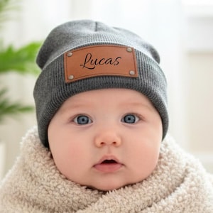 Puede incluir: Un bebé con un gorro gris con un parche de cuero marrón que dice "Lucas". El bebé está envuelto en una manta color crema y sentado en una cesta de mimbre blanca. El bebé tiene ojos azules.