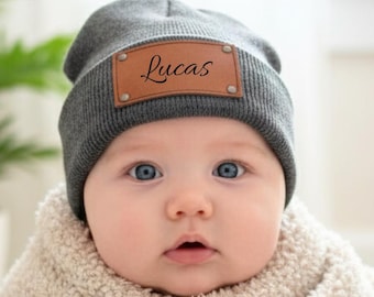 May include: A baby wearing a grey beanie hat with a brown leather patch that reads "Lucas". The baby is wrapped in a cream-coloured blanket and sitting in a white wicker basket. The baby has blue eyes.