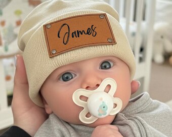 May include: A baby wearing a beige knit beanie with a brown leather patch that reads "James" in cursive. The baby is also wearing a gray onesie and a pacifier. The background includes a crib and a teddy bear.