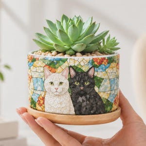 Puede incluir: Una maceta cilíndrica de cerámica con un colorido patrón de mosaico de un gato blanco y un gato negro. La maceta contiene una planta suculenta verde y se asienta sobre un platillo de madera circular. Los gatos tienen texturas de pelaje detalladas.