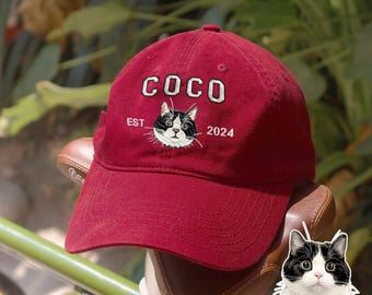 May include: A deep red baseball cap featuring white embroidered text that reads COCO, EST, and 2024. Centered between the text is a detailed embroidered graphic of a black and white cat face with large eyes, matching the cat shown below the cap.