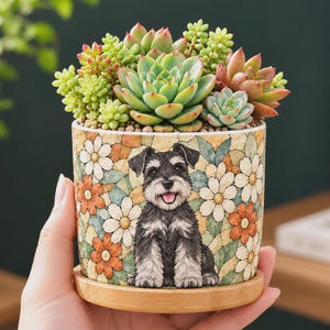 May include: A cylindrical ceramic planter featuring a mosaic-style pattern of white and orange daisies with green leaves. A black and gray Schnauzer dog illustration is centered on the front. The planter sits on a light wood saucer.