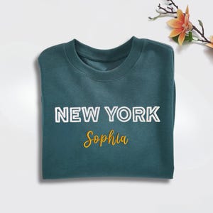 May include: A folded teal sweatshirt with the words "NEW YORK" in white block letters and the name "Sophia" in yellow script. The sweatshirt is made of a soft material and is folded neatly.