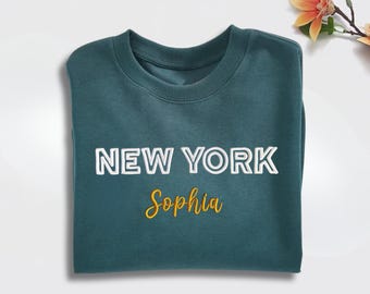 May include: A teal sweatshirt folded neatly, featuring the text "NEW YORK" in white and the name "Sophia" in yellow script. The garment is made of a soft material, perfect for casual wear.