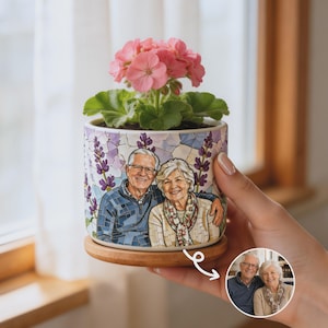 May include: A cylindrical ceramic planter with a wooden base, featuring a mosaic-style illustration of an older couple. The planter holds a pink geranium plant. A small circular inset shows the original photograph of the couple used for the design.