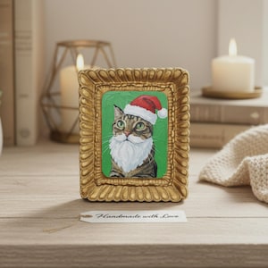 Pode incluir: Pintura emoldurada de um gato usando um gorro de Papai Noel e barba branca em um fundo verde. A moldura é dourada e ornamentada. Uma etiqueta abaixo diz "Handmade with Love".