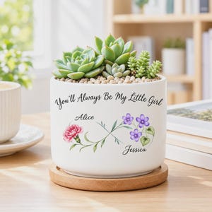 May include: A white cylindrical ceramic planter containing various green succulents. The planter features the black text "You'll Always Be My Little Girl" above an infinity symbol graphic with a pink carnation, purple flowers, and the names Alice and Jessica.