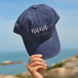 Może przedstawiać: Granatowa czapka bejsbolówka z białym haftem z napisem "nana". Pod spodem znajduje się napis "EST.2025". Czapka ma zakrzywiony daszek i guzik na górze.