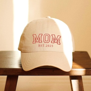 May include: A beige baseball cap with the word "MOM" in red block letters and "EST.2025" printed below. The cap is sitting on a wooden surface. The hat is a casual accessory.