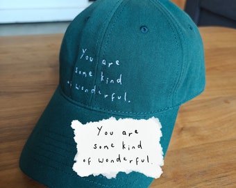 Puede incluir: Una gorra de béisbol verde con la frase "You are some kind of wonderful." bordada en la parte delantera. Una nota escrita a mano con la misma frase está unida a la visera.