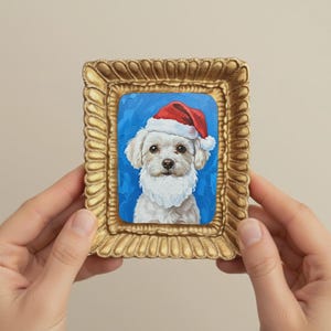 Pode incluir: Pintura emoldurada de um cão branco usando um gorro e barba de Papai Noel contra um fundo azul. A moldura dourada tem um design ornamentado. Os olhos do cão são escuros e a pintura tem uma aparência texturizada.