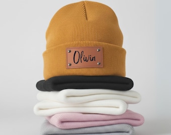 May include: Mustard yellow beanie with a brown leather patch displaying the name "Olivia" in black script. Stack of folded beanies in black, white, pink, and grey below. The beanies are made of a soft knit material.