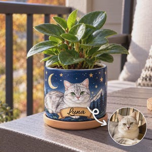 May include: A cylindrical dark blue ceramic planter featuring a graphic illustration of a silver tabby cat, a crescent moon, stars, and mountains. A banner with the name Luna is printed on the front. A green plant sits inside the planter, which rests on a light brown wooden saucer. A circular inset shows the original cat photo.