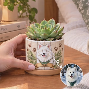 May include: A cylindrical ceramic planter featuring a light wood grain pattern, brown paw prints, and a green leafy border. The front displays a custom illustration of a white Samoyed dog wearing a green harness with the name Bella printed below.
